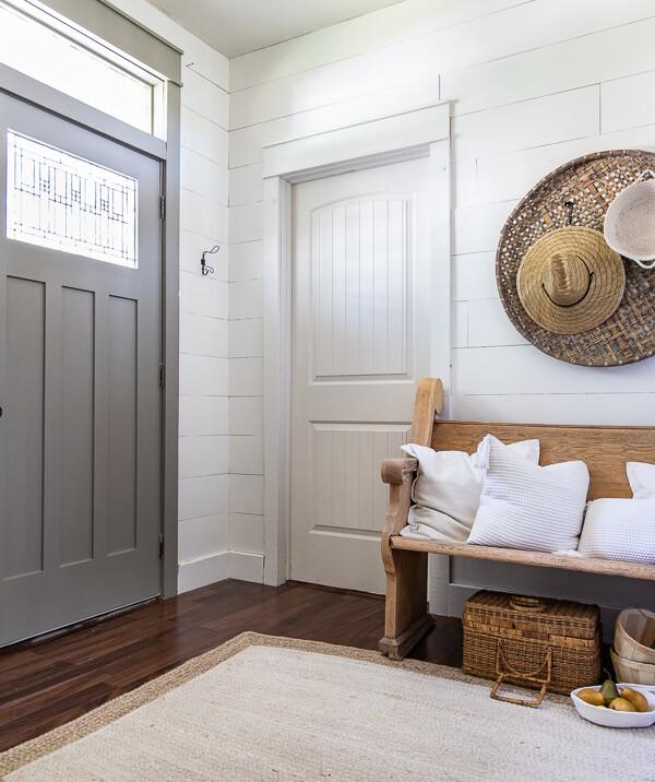 Door painted with Benjamin Moore Gettysburg Grey. Fall decor ideas including my wooden church pew in the entry with baskets on the wall, topped with straw hats and rope baskets. White pillows top of the vintage church pew and a basket of pears adds a touch of color to this neutral fall entryway.
